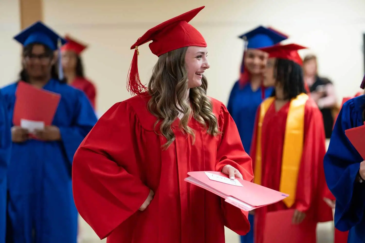 Una joven con toga y birrete rojos sonríe con una carpeta de diplomas en la mano.