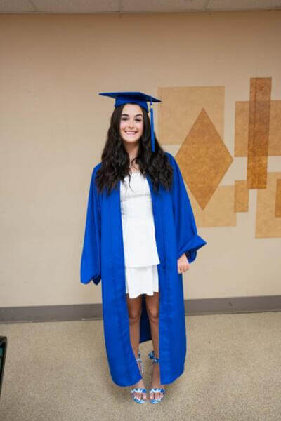 Una joven sonríe, con toga y birrete azul de graduación sobre un vestido blanco.