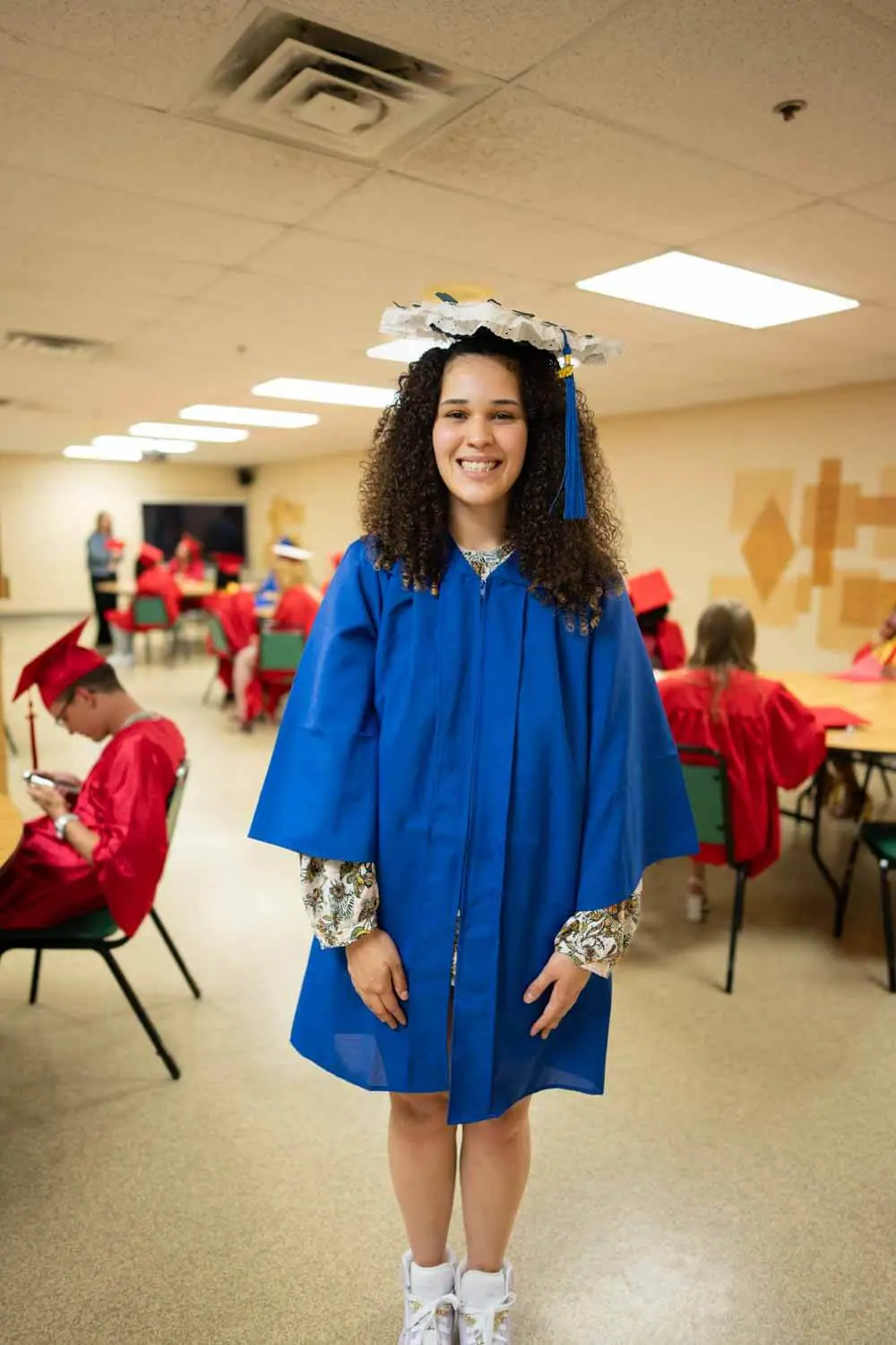 Una joven sonriente con toga azul de graduación y birrete decorado en un aula.