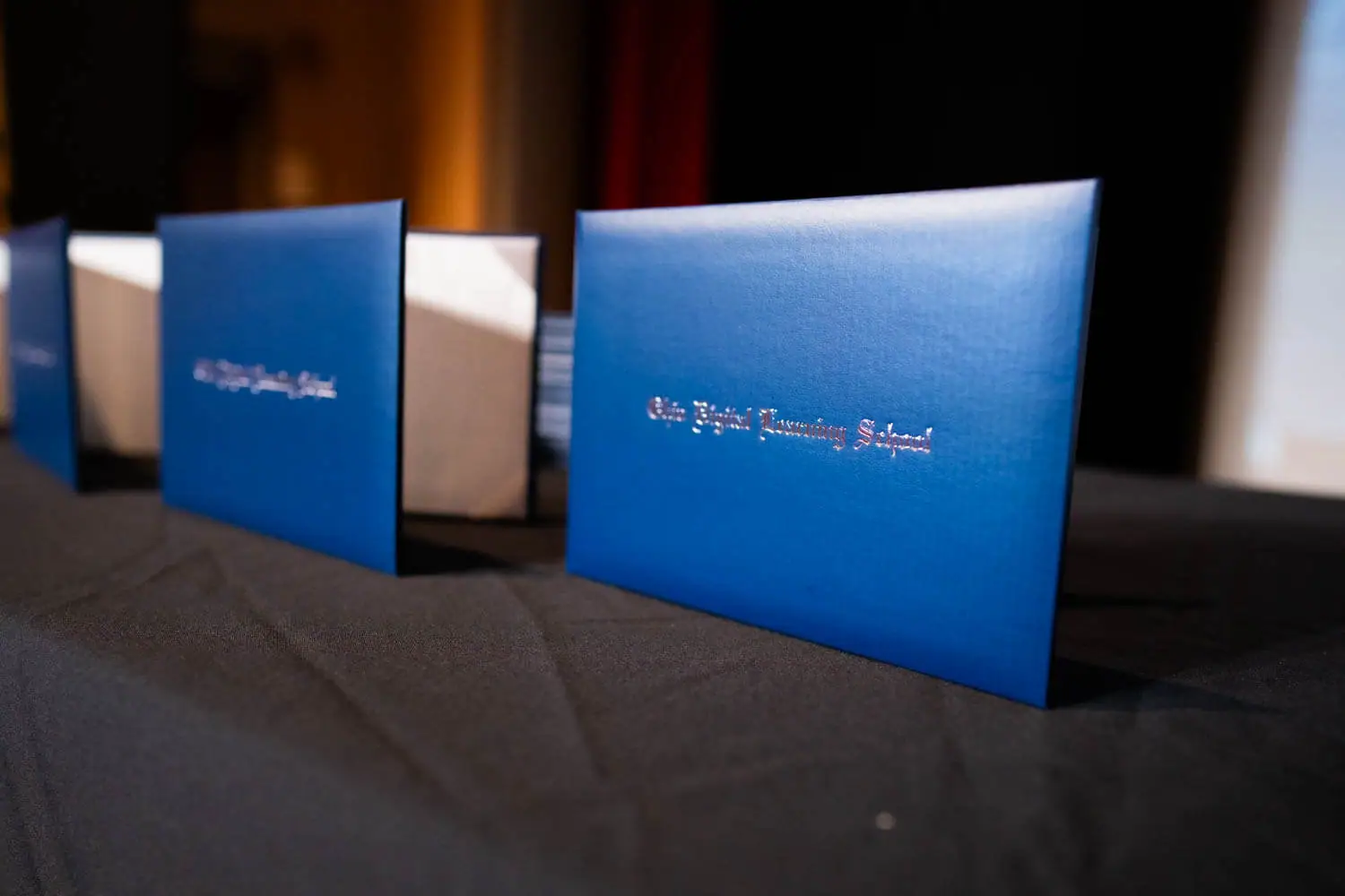 Diplomas azules alineados sobre una mesa negra, reflejando la luz ambiente.