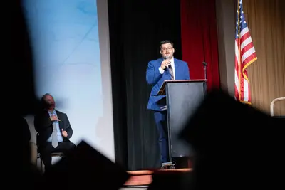 Un hombre con traje azul habla en un podio en el escenario con una bandera estadounidense a su lado.