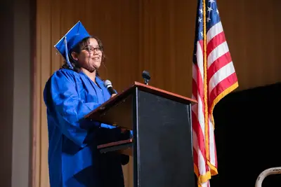Un graduado con toga y birrete azul habla con confianza en un podio con micrófono.
