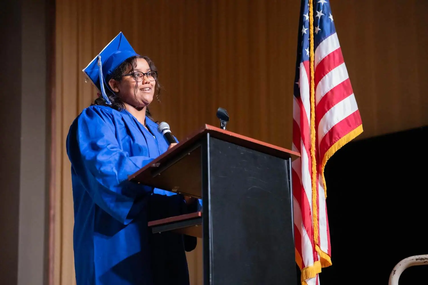 Un graduado con toga y birrete azul habla con confianza en un podio con micrófono.