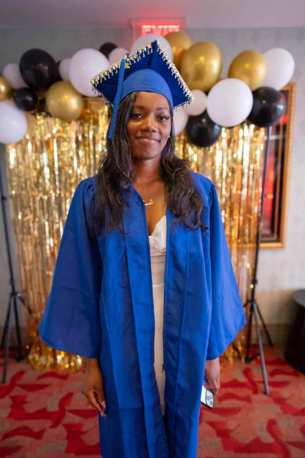 Una joven con toga y birrete de graduación azul sonríe delante de unos globos dorados y negros y un fondo brillante.