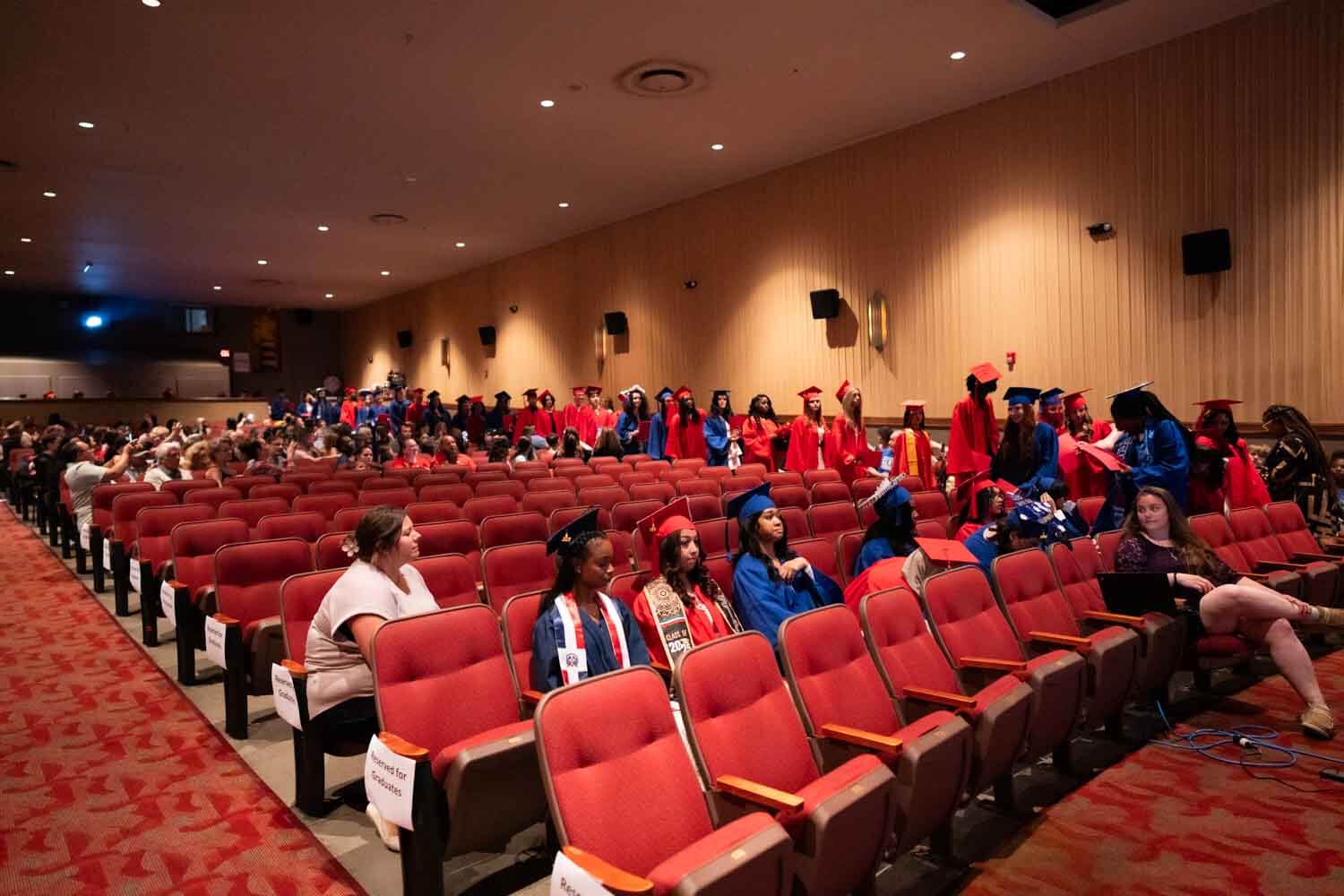 Los graduados con togas y birretes rojos y azules llenan los asientos del auditorio, algunos de pie y charlando.