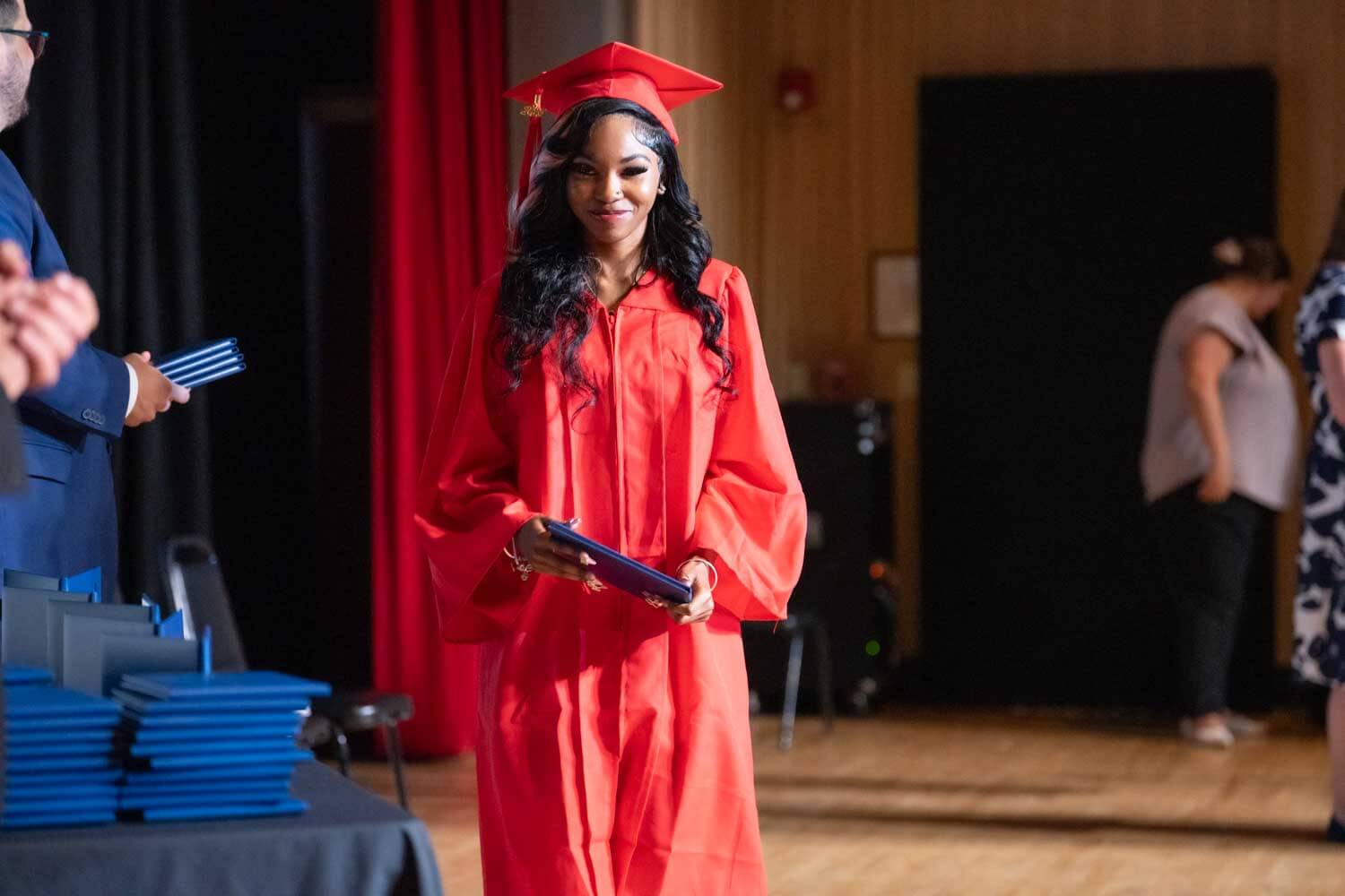 Un graduado con toga y birrete rojo sonríe mientras sostiene un diploma en el escenario.