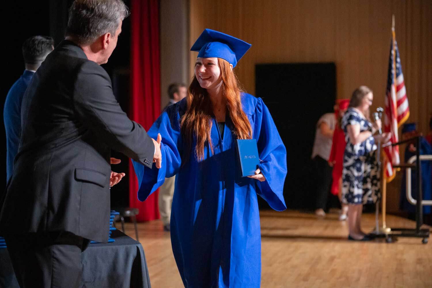 Una joven con toga y birrete azul sonríe mientras estrecha la mano de un hombre en el escenario de una ceremonia de graduación.