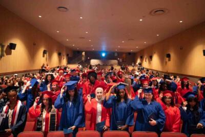 Graduados con togas y birretes rojos y azules llenan un auditorio, ajustándose las borlas.