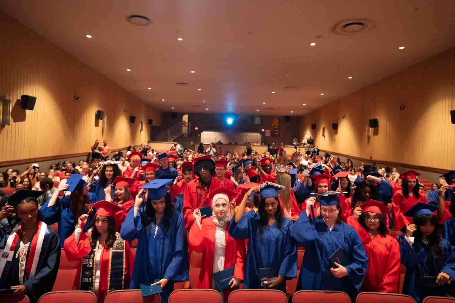 Graduados con togas y birretes rojos y azules llenan un auditorio, ajustándose las borlas.