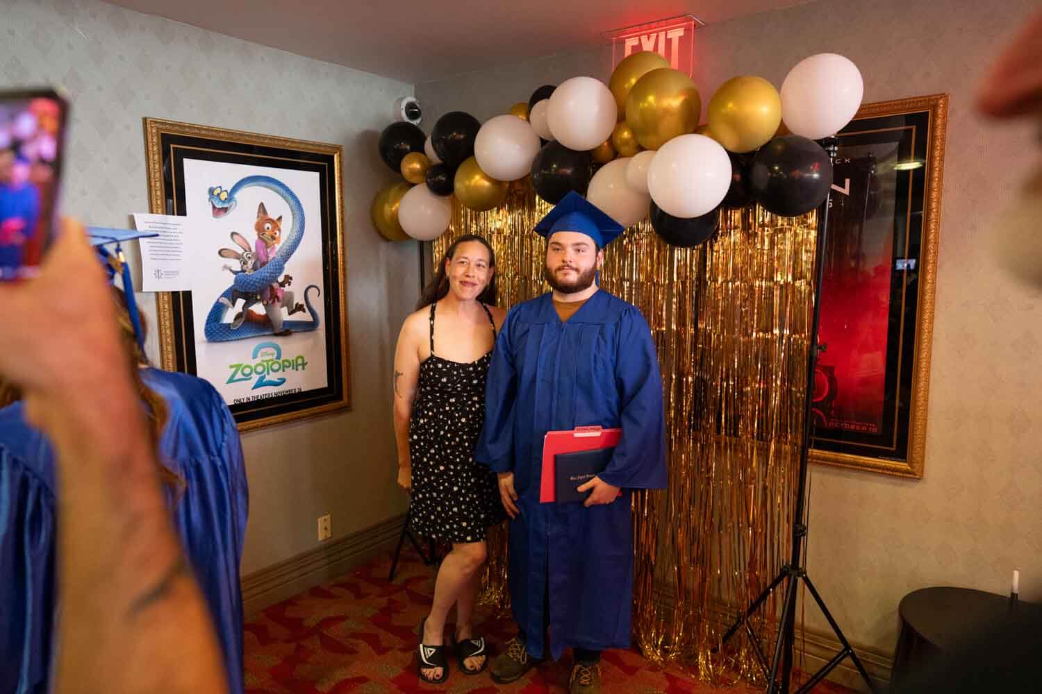 Un graduado con toga y birrete azul posa con una mujer vestida de negro.