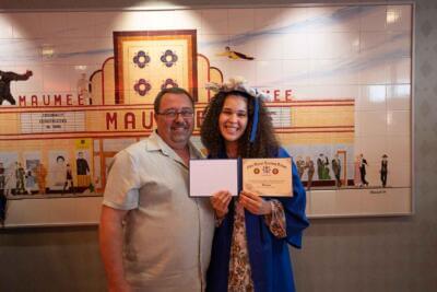 Una joven sonriente, con toga y birrete azules, sostiene un diploma junto a un hombre.