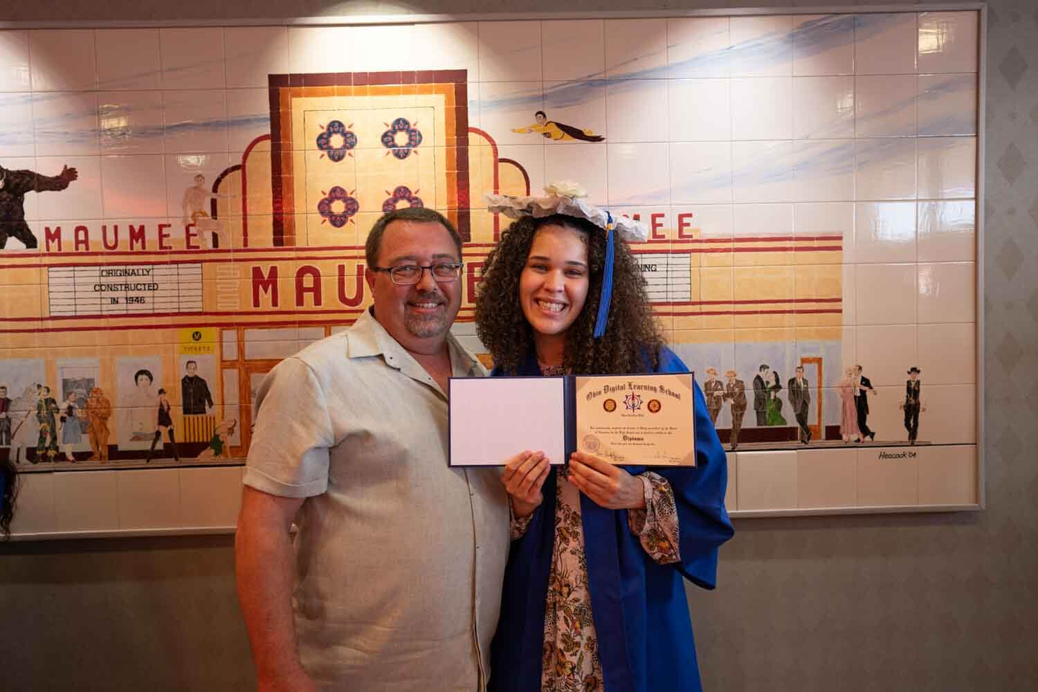 Una joven sonriente, con toga y birrete azules, sostiene un diploma junto a un hombre.