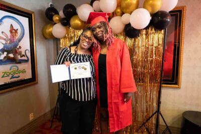 Un graduado con toga y birrete rojos posa con una mujer que sostiene un certificado.