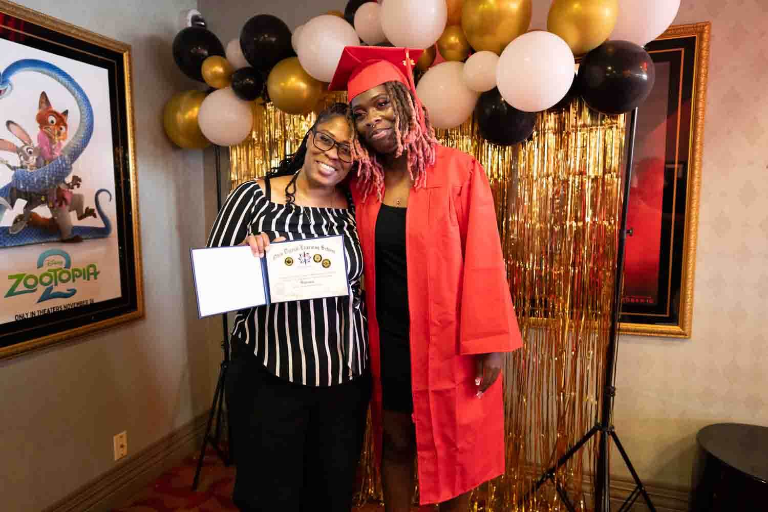 Un graduado con toga y birrete rojos posa con una mujer que sostiene un certificado.