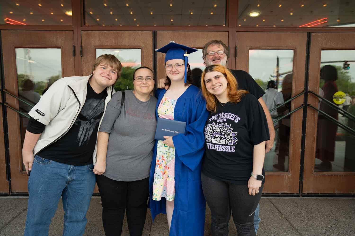 Un graduado con toga y birrete azul sostiene un diploma, rodeado de cuatro personas sonrientes.