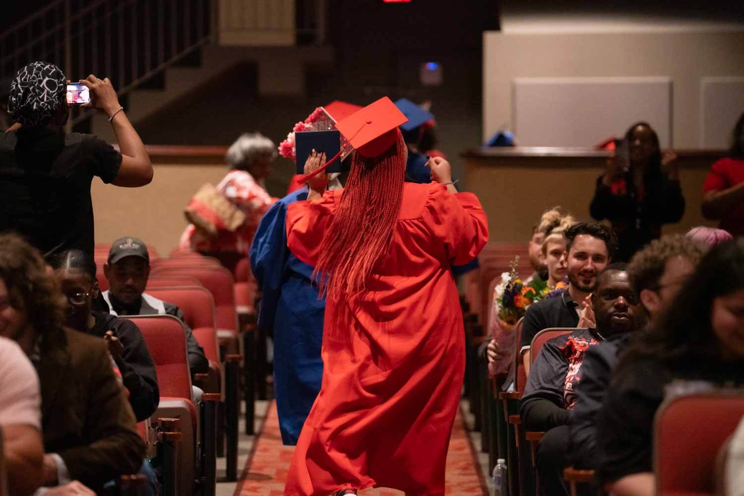 Un graduado con toga y birrete rojos baila alegremente por el pasillo de un auditorio.