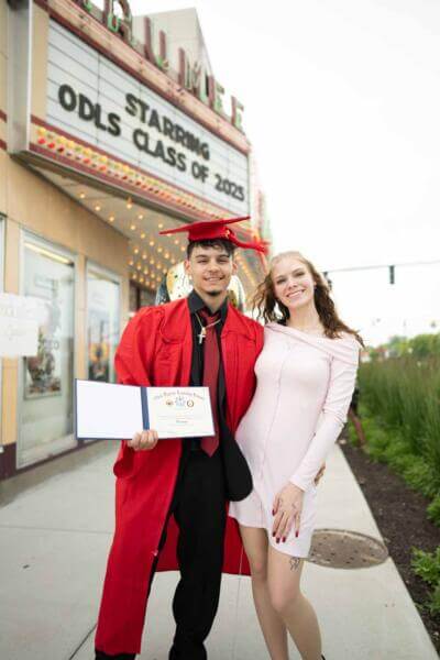 Un graduado con toga y birrete rojos sonríe con su diploma junto a una mujer con un vestido claro.