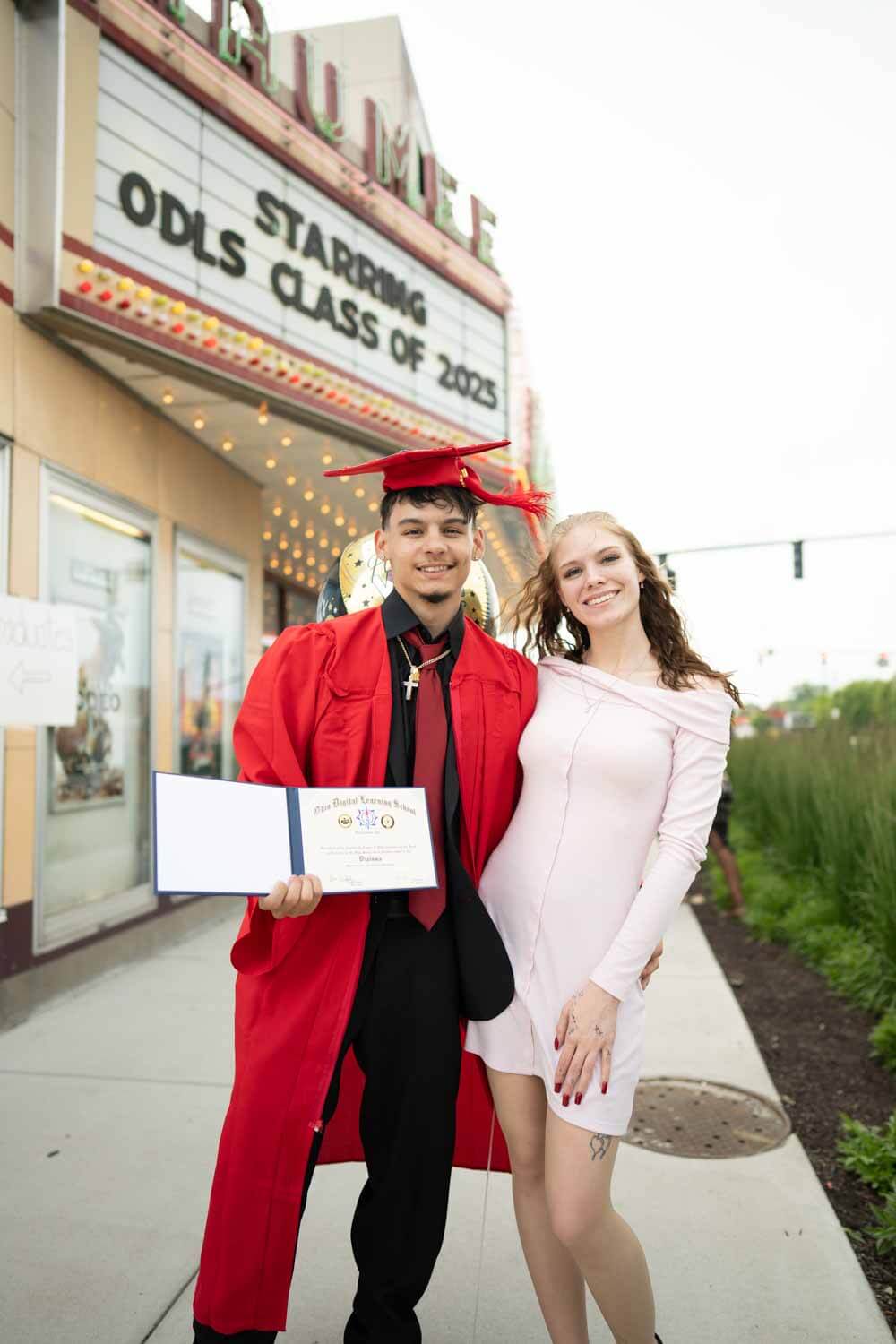 Un graduado con toga y birrete rojos sonríe con su diploma junto a una mujer con un vestido claro.