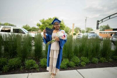 Una graduada sonriente con toga y birrete azul sostiene un diploma y un ramo de flores.