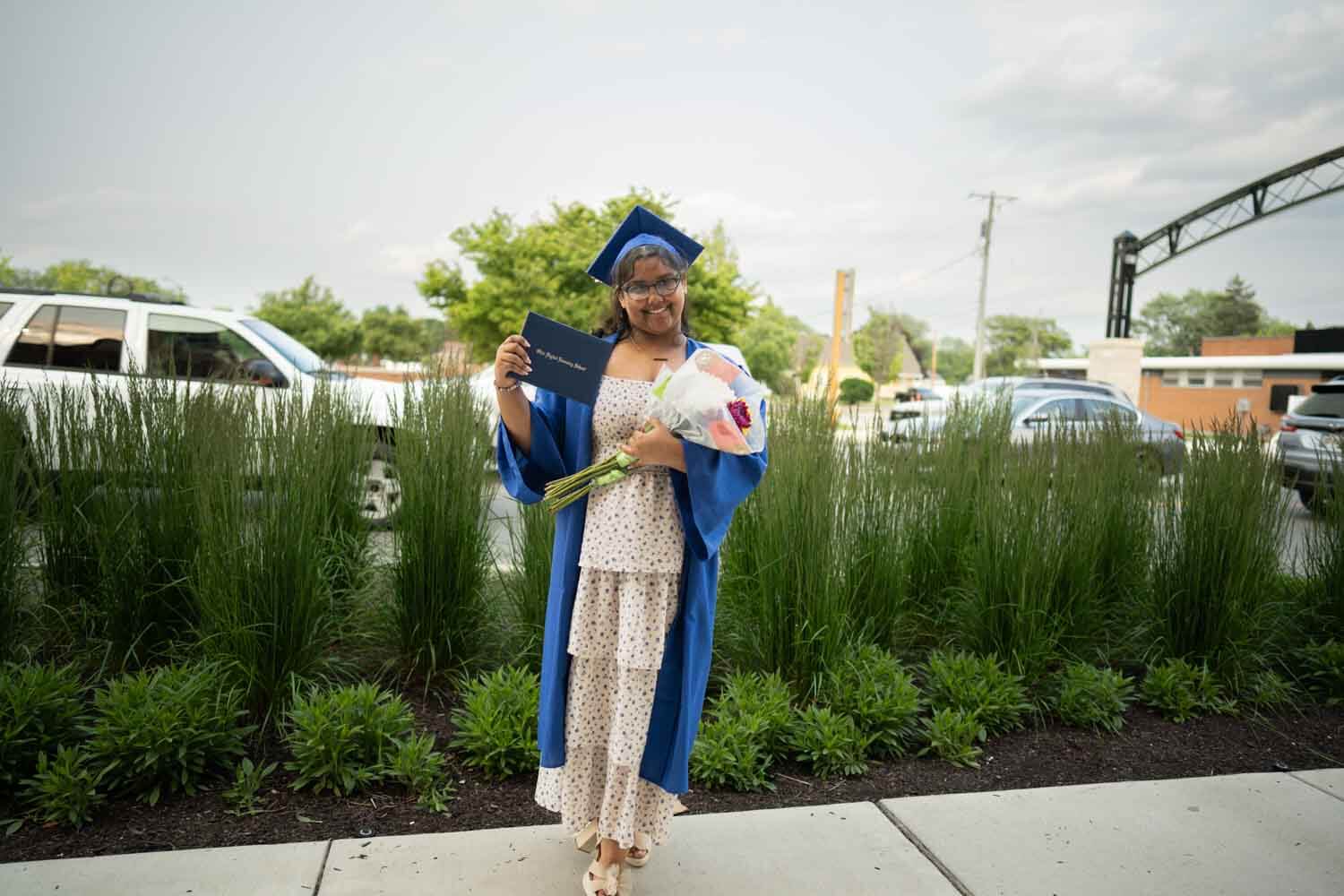 Una graduada sonriente con toga y birrete azul sostiene un diploma y un ramo de flores.